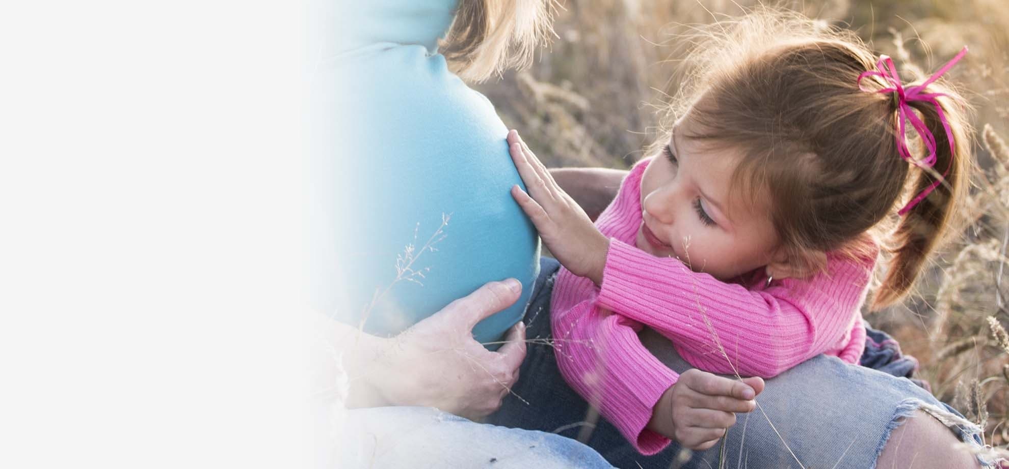 A young girl in a pink sweater resting her hands and cheek on her pregnant mother's belly in a sunlit field
