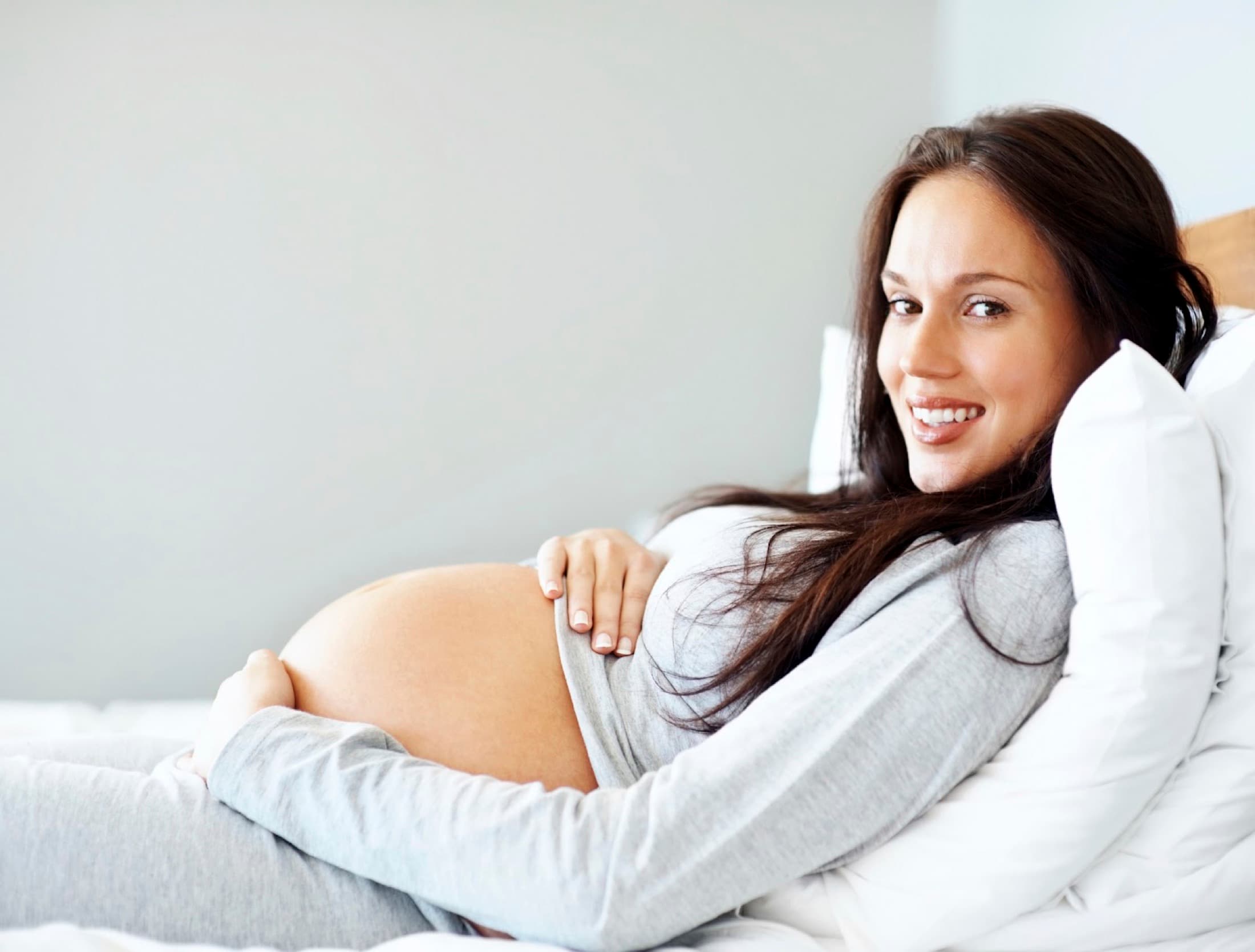 A pregnant woman resting against white pillows, smiling with both hands on her belly